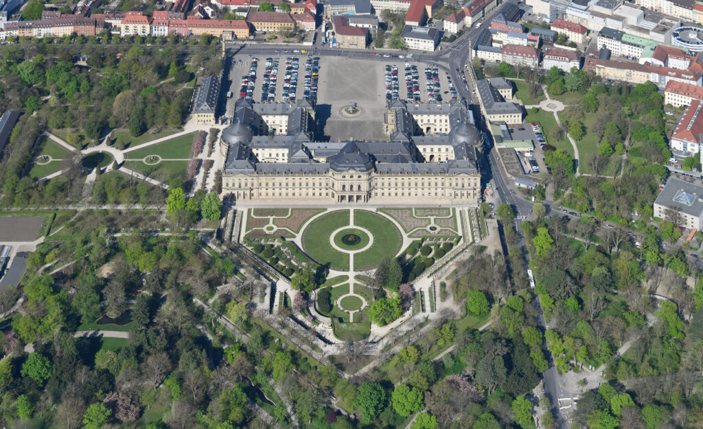 Aerial view of grand palace with formal gardens and symmetrical courtyards surrounded by urban development