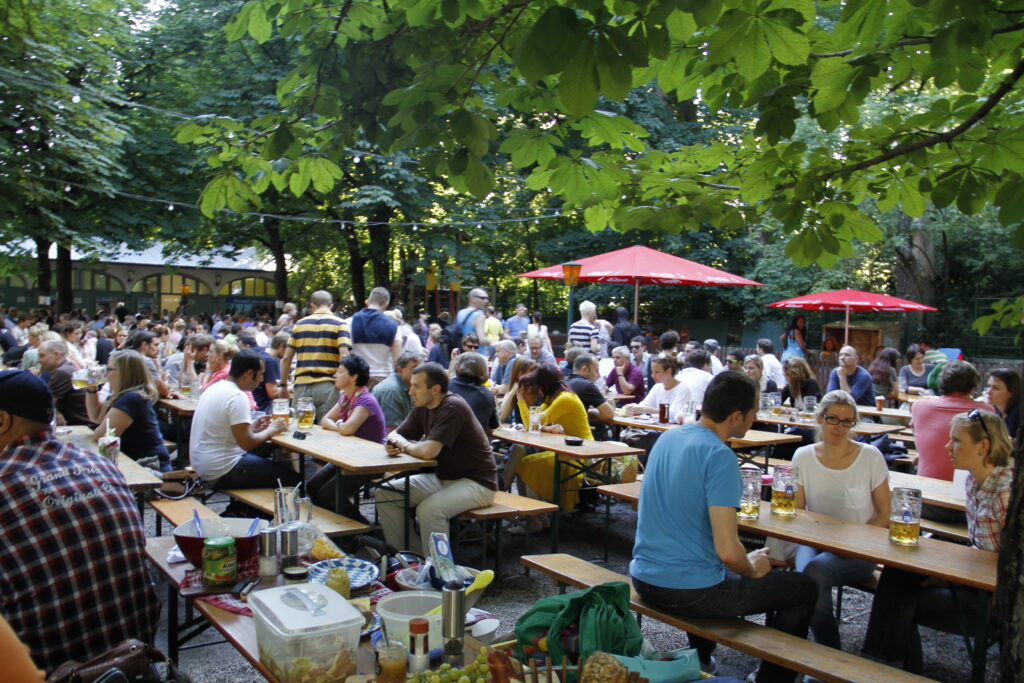 Crowded outdoor beer garden with long tables and red umbrellas under trees