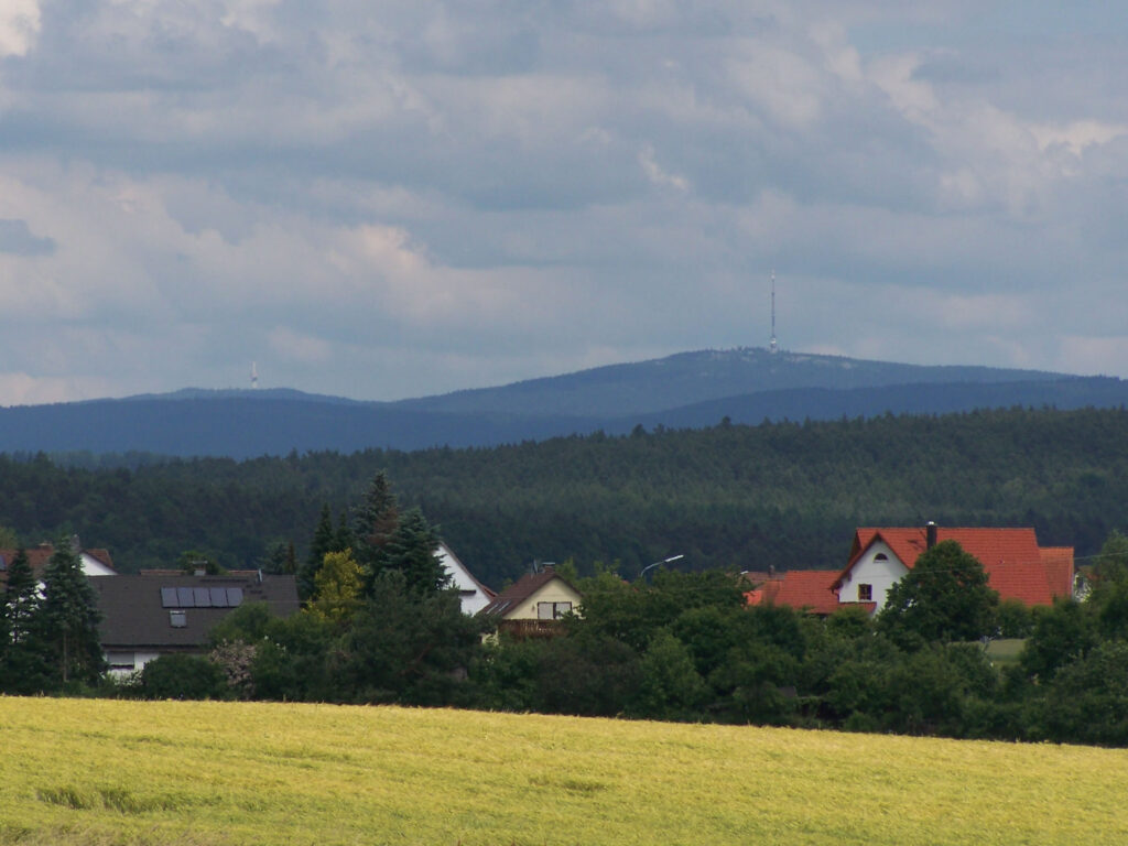 Rural German valley with forested hills, red-roofed houses, and distant communication towers