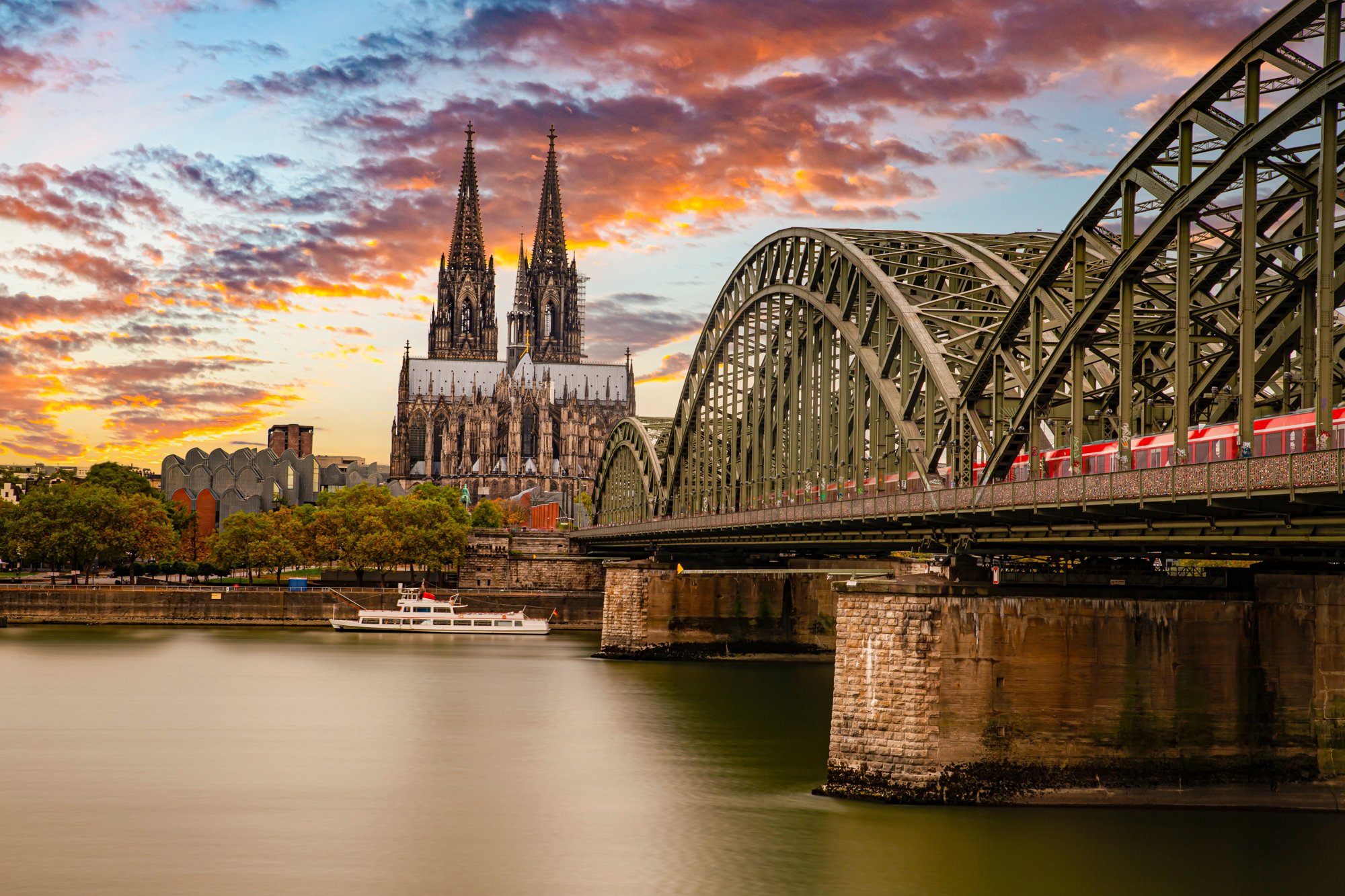 Red train crossing the Hohenzollern Bridge over the Rhine with Cologne Cathedral behind at sunset